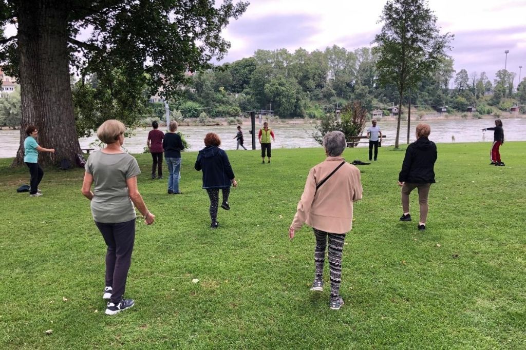 Eine Gruppe von sportlichen Seniorinnen folgt in einem Park den Anweisungen zweier Kursleitenden.