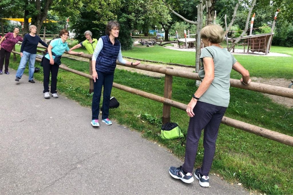 Seniorinnen halten sich an Zaun fest und führen mithilfe ihrer Kursleiterin sportliche Übungen aus.