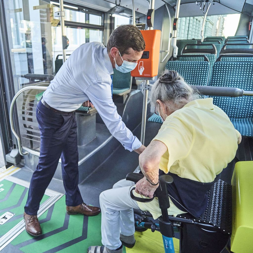Mitarbeiter hilft sitzenden Seniorin im Bus bei der Befestigung ihres Rollators.