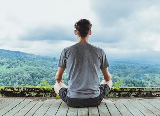 Meditierender Mann sitzt auf einer Holzterrasse und hat einen Ausblick auf einen grossen Wald.