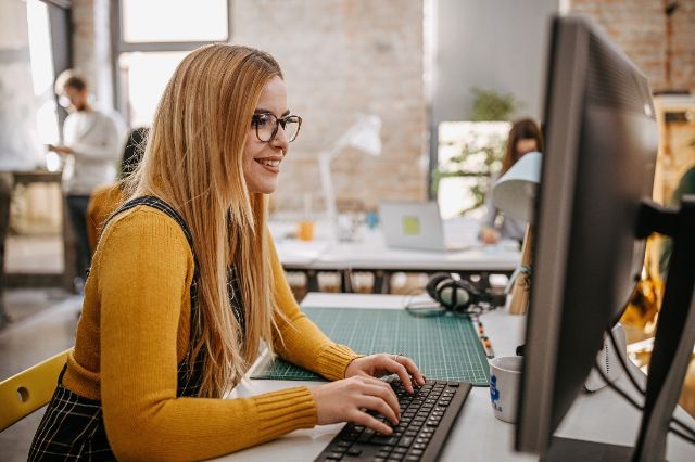 Eine junge blonde Frau arbeitet als Assistentin am Computer in einem grossen Büro.