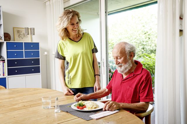 Ein Senior sitzt am Tisch und freut sich auf das Mittagessen, welches ihm eine Mitarbeiterin nach Hause gebracht hat.