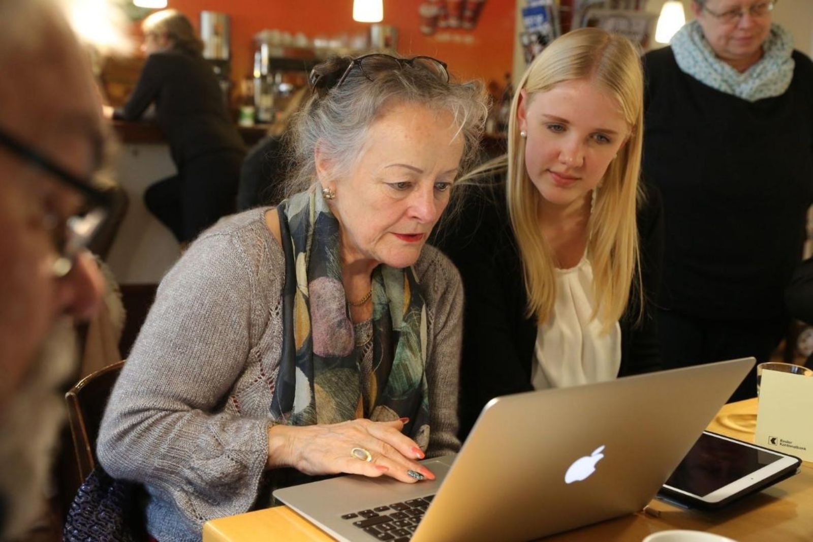 Junge Mitarbeiterin hilft Seniorin im Digital Café mit ihrem Laptop und iPad.
